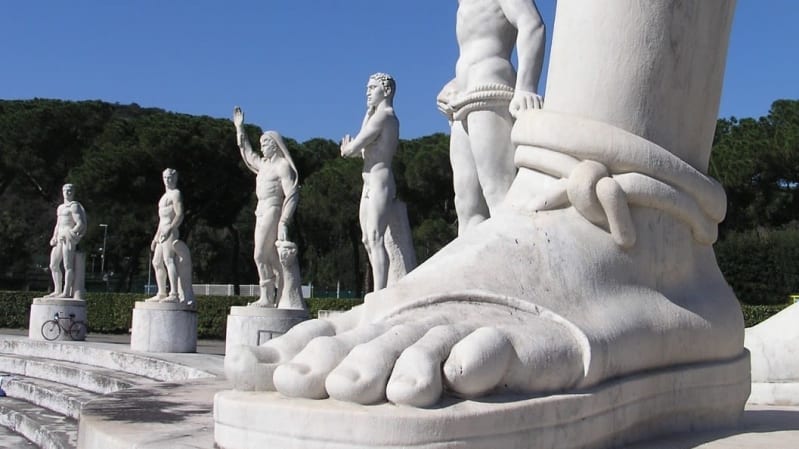 Large white marble statue in the foreground, only the sandal is visible. Behind in a row are five other white marble statues of naked and semi-naked people.