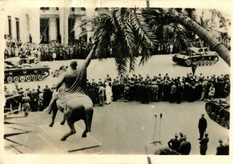 Black and white photo of a statue on a plinth, which is a man on horseback giving a salute. Tanks and rows of people watching in the background. A large palm tree is to the right in the foreground.