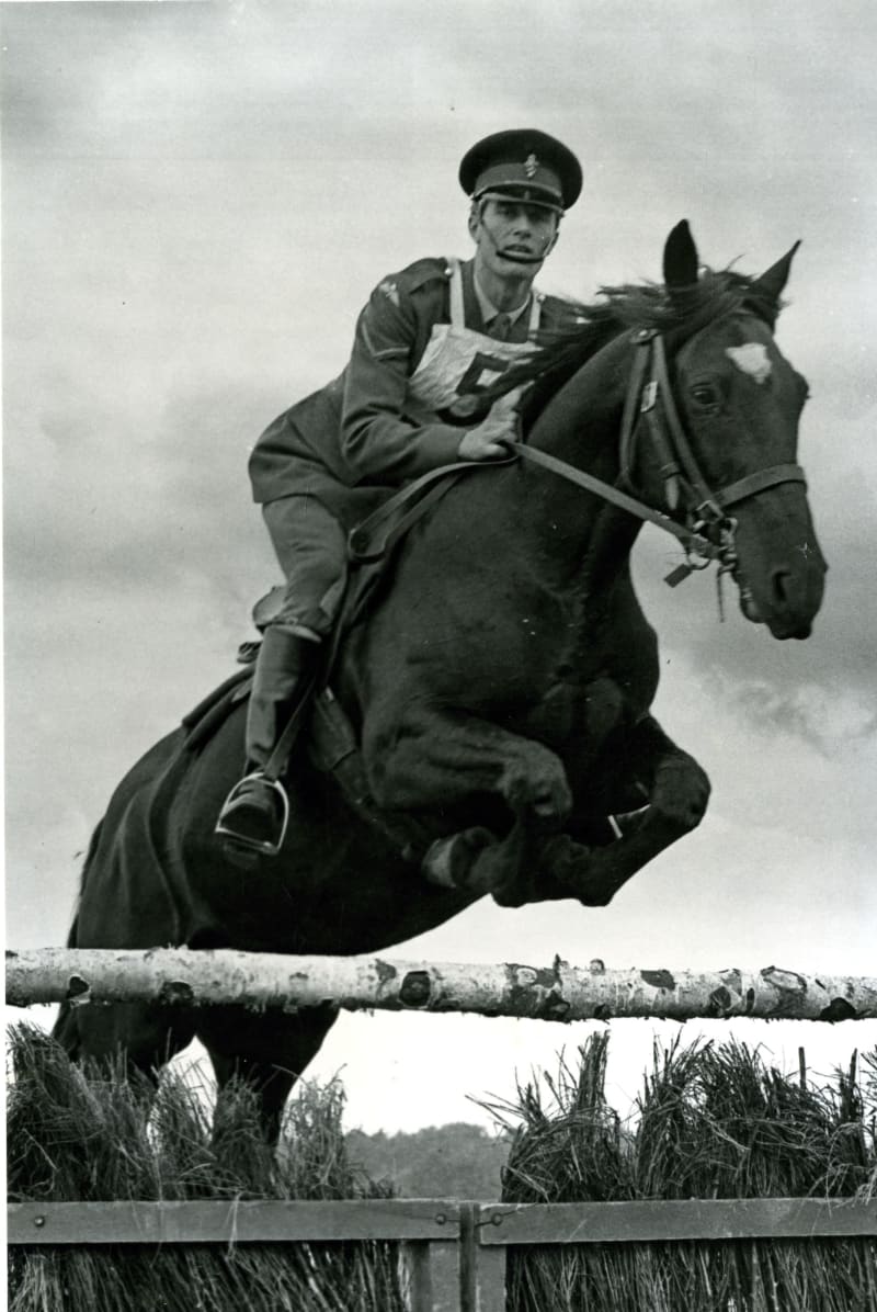 A man in army dress uniform and hat riding a horse which is jumping over a fence. The man wears a bib with the number 5.