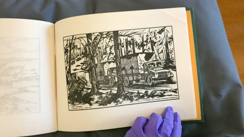 Landscape sketchbook open on a page with a cartoon sketch of military trucks driving through a forest. A purple gloved hand holds the book.