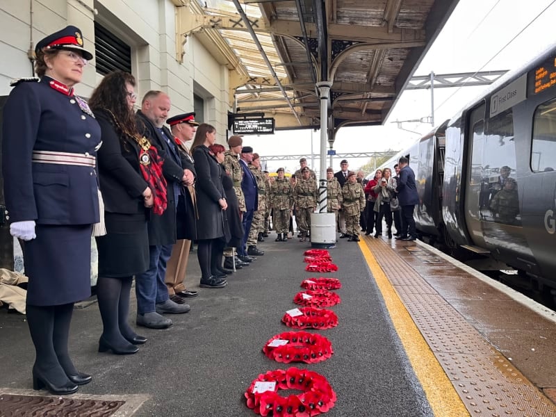 People stood in a line on a train platform with poppy wreaths laid in a line in front of them.