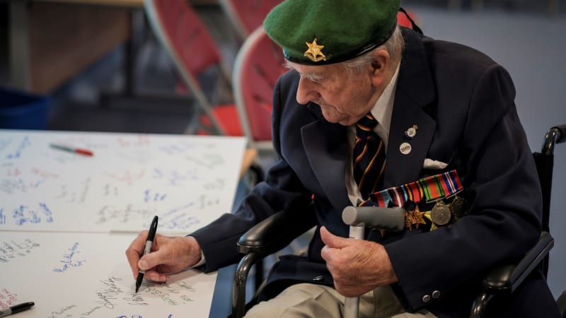 Veteran wearing medals sits in wheelchair and signs large piece of paper on table. The paper has multiple signatures and messages.