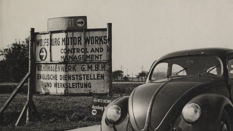 Black and white image of VW Beetle in foreground and sign with English and German writing in background. English writing at the top of sign reads: " Wolfsburg Motor Works. Control and Management ". 
