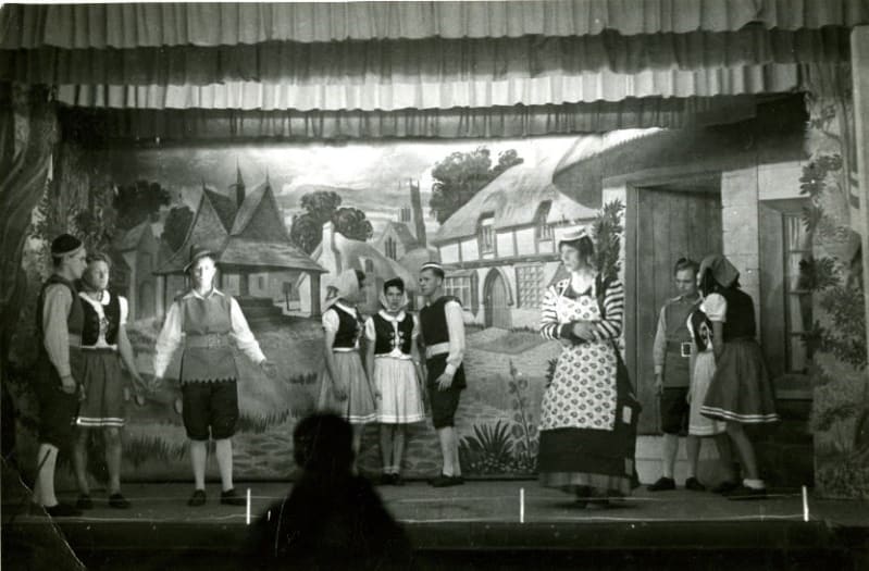 Black and white photo of nine actors on a stage dressed in costume. The set is painted like a village.