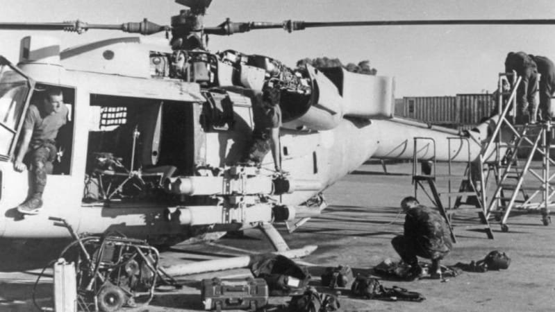 Black and white photo of men in military uniforms fixing a helicopter. Two men sit on helicopter, one crouches down on the ground and two more stand at the top of a ladder. Mechanical equipment is on the ground.