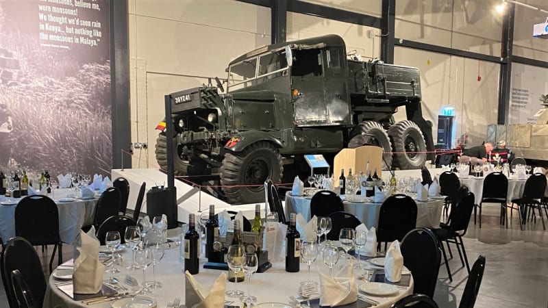 Tables set up for dinner with tablecloths and china, inside a large hangar, a dark green vehicle in the background.