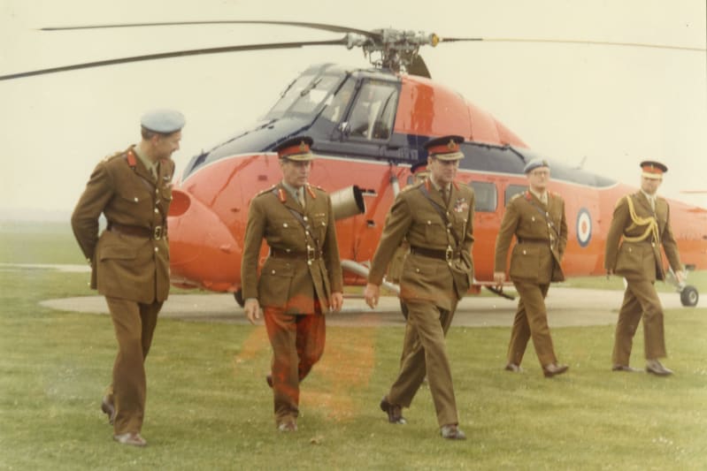 Prince Philip walks amongst four other soldiers outside on grass. All wear uniform. In the background is a red and blue helicopter on the ground. 