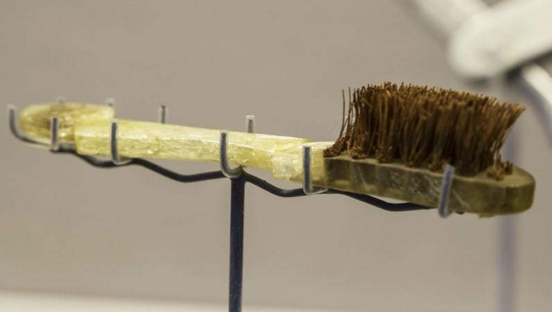 Old yellow toothbrush with dark brown bristles being held up sideways on a stand. 