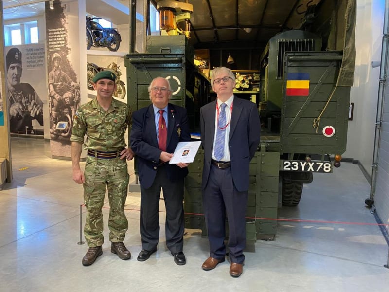 Three men (one in camouflage, two in suits) stand in front of a vehicle in a museum. The centremost man holds a book.