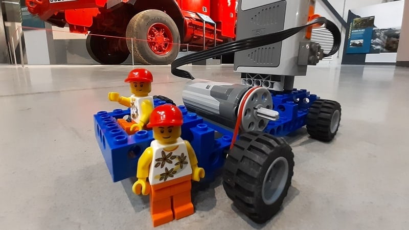 A LEGO car with two LEGO people and a motor on top. Positioned on the floor inside a vehicle gallery, a large red vehicle visible in the background.