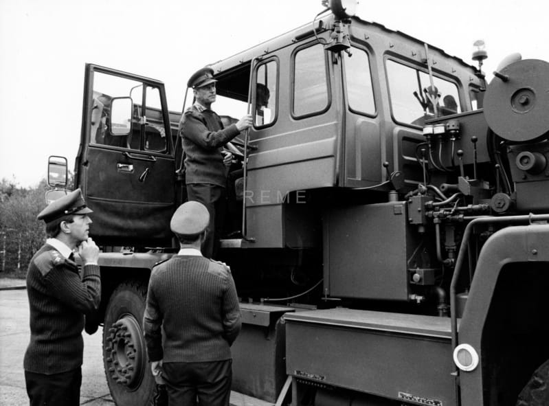 Black and white photograph of Prince Philip standing in the doorway of a large military vehicle. He looks out towards the back of the vehicle. Two other soldiers stand on the floor beneath and look at the vehicle.