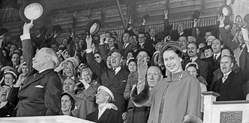 Black and white image of the Queen smiling and waving, a crowd is seen doing the same in the background.