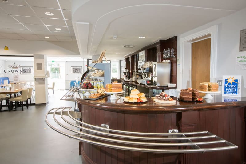 A brown cafe counter with cakes and scones on top. Coffee bar and drinks behind. Restaurant seating in background.