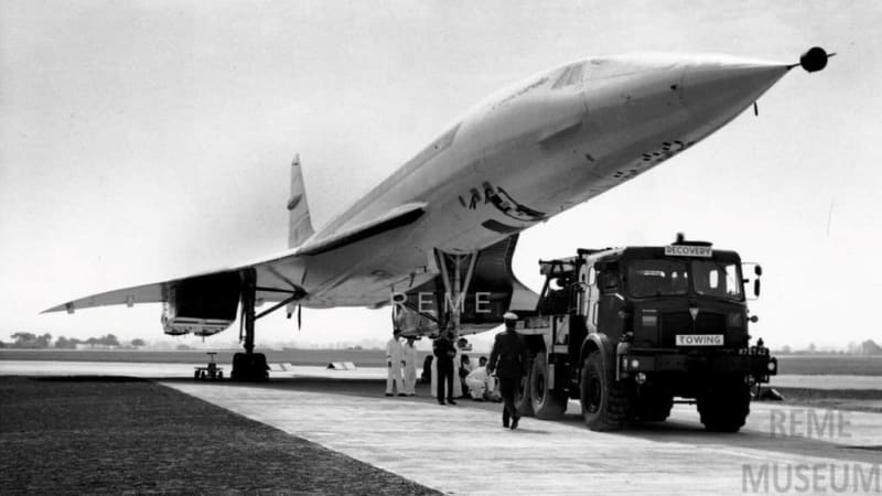 Black and white photo of large plane on runway with people underneath. A military vehicle is parked in front.