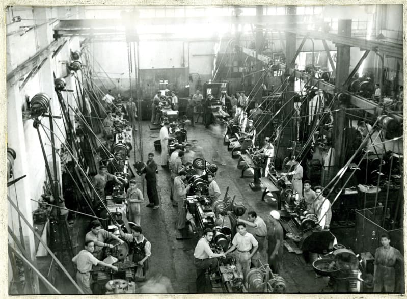 Black and white photo of a busy workshop. There are three long rows of stations, and pulleys attached to them from the ceiling. Men work with machines at the stations.
