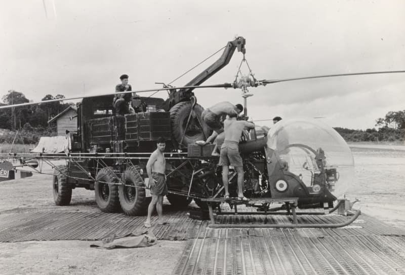 Black and white photo of a large military truck carrying a helicopter. A man sits on top of the truck, another man stands next to it, and three more men are fixing the helicopter.