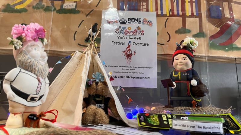 Small display with two gnomes painted in clothes, one wearing a flower crown, tent teepee, colourful fairy lights and a poster titled Festival Overture.