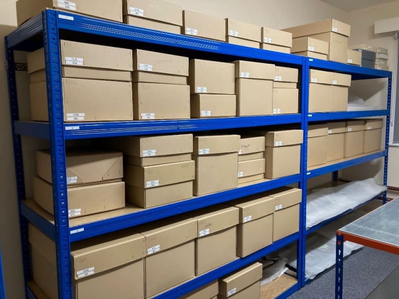 Blue metal storage shelves filled with brown cardboard boxes in a room. The boxes have small white labels on them.