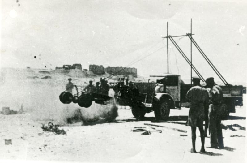 Black and white image. A truck pictured in the desert with attachment to the front creating a cloud of dust, soldiers stand nearby watching on.