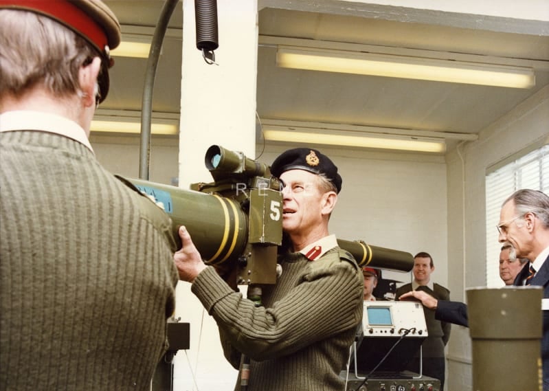 Prince Philip holds a large tank gun and looks through the gunsight. Other men stand nearby. 
