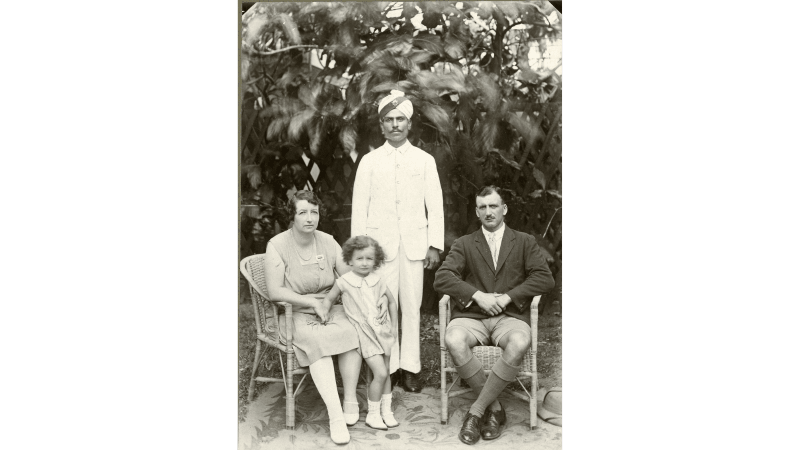 Rowcroft and his wife sit in wicker chairs, their child stands in between. Behind stands an Indian man dressed in white with a turban 