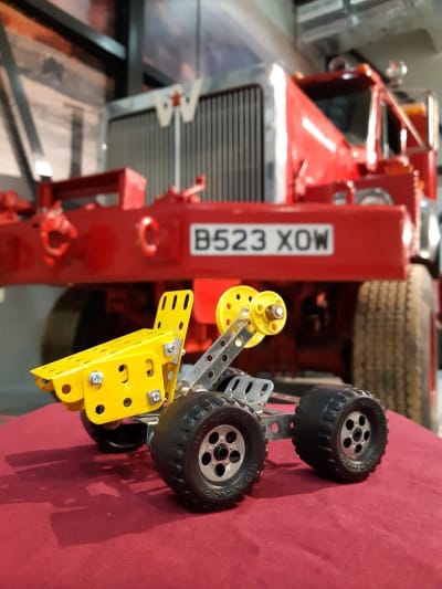A yellow Meccano built vehicle sitting on a red table with a large red vehicle in the background, which is slightly out of focus.
