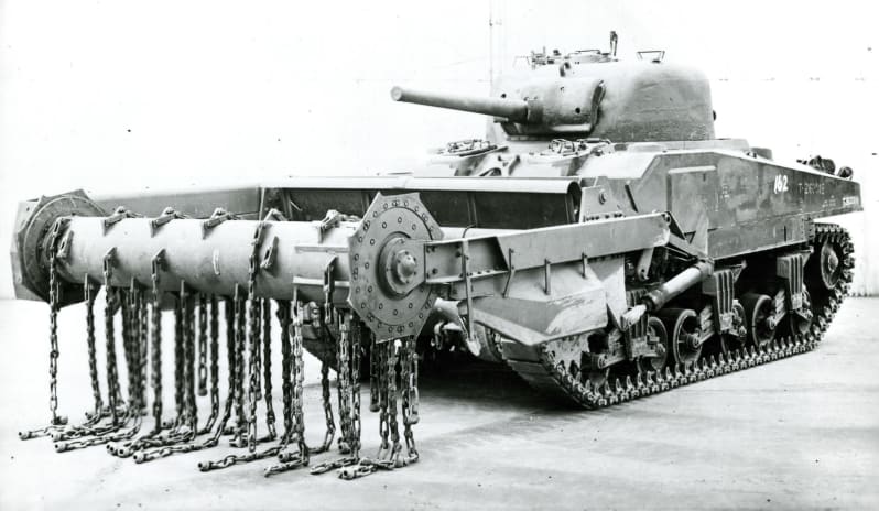 Black and white photo of a tank with long arms out front with roller attached, to which chains are attached and hanging down. 