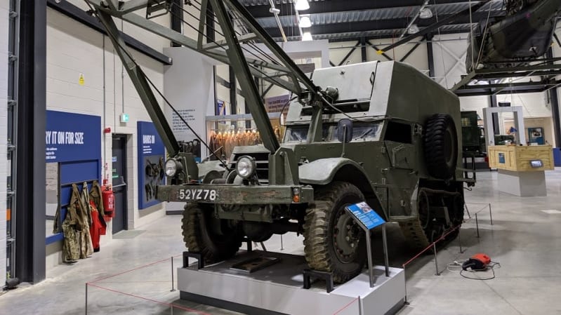 A vehicle with both wheels and tracks and a crane on the front, situated inside a large hangar with other displays around it.