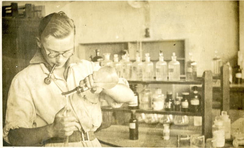 Black and white photo of a man smoking a pipe and pouring liquid from a balloon flask into a test tube. Labeled bottles of liquid on shelves behind him in background.