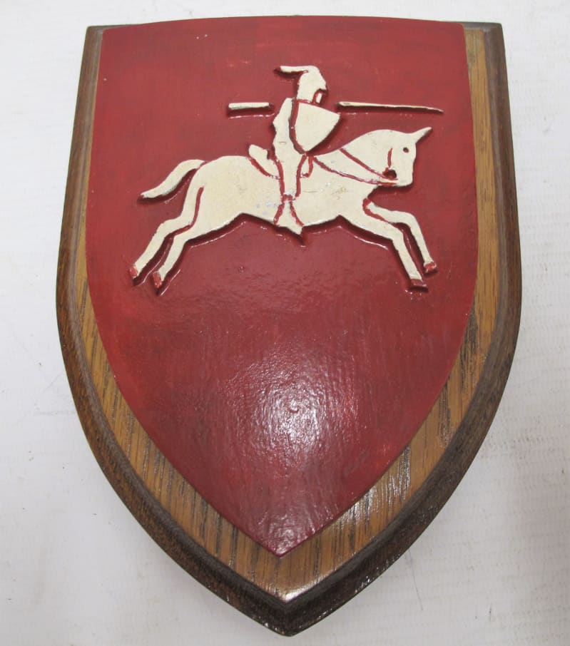 A wooden shield shaped plaque with red painted top. Carved into the centre is a white unicorn (pegasus) ridden by a knight.