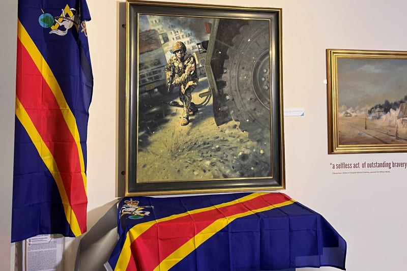 Painting with display case below covered with a blue red and yellow REME flag, another flag hung over a panel on the wall.