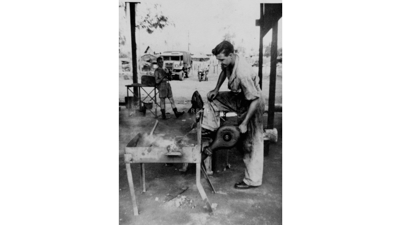 Black and white photograph shows a man operating machinery outside. Another man stands in the background, a vehicle is further behind.