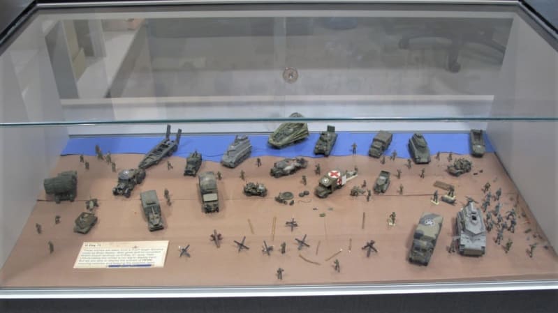 Birds eye view of scale model vehicles and people laid out in a diorama scene inside a display case.
