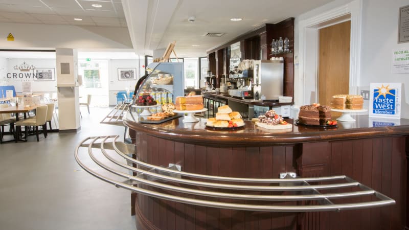 A brown cafe counter with cakes and scones on top. Coffee bar and drinks behind. Restaurant seating in background. 