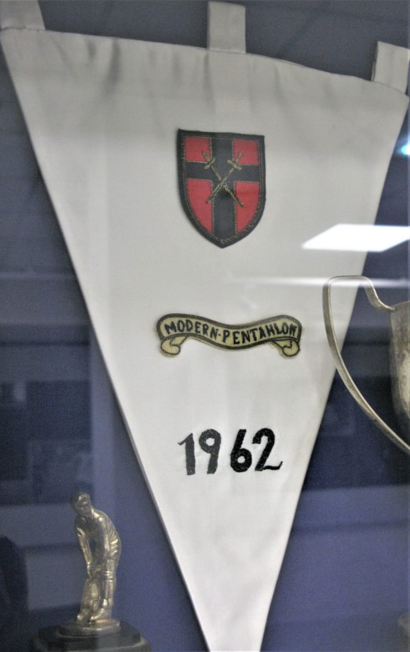 White satin triangular pennant with point downwards, three loops at the top for hanging. Embroidered onto the front is a shield, red and navy, a banner reading Modern Pentathlon and 1962.