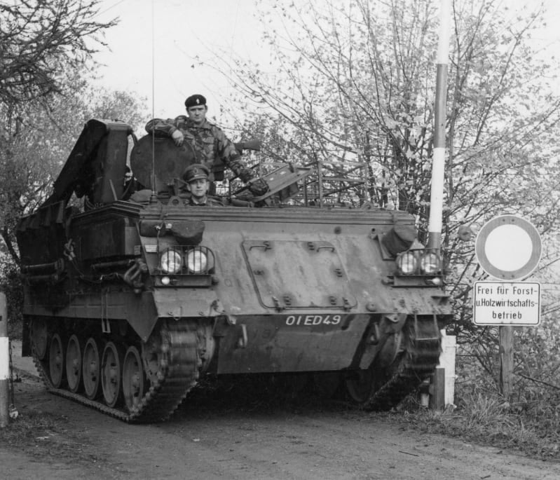 Black and white photograph of Prince Philip and another soldier sitting in a tank travelling past an opened barrier. Only their torsos are visible. Trees and a circular sign with German writing udnerneath at the side of the path.