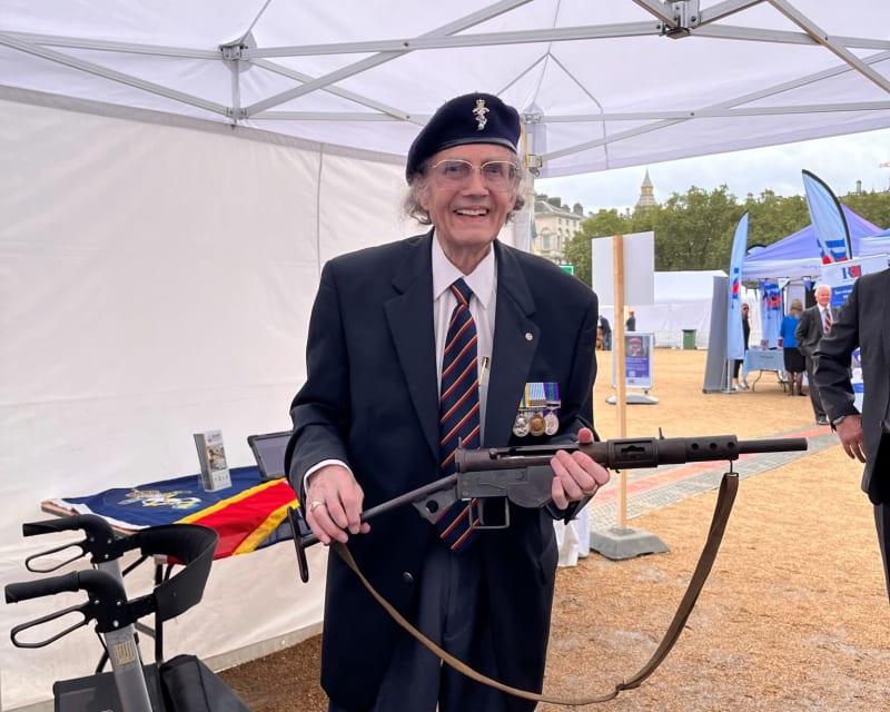 A man in a suit and REME beret stands inside a white outdoor gazebo, smiling and holding a metal gun in both hands.