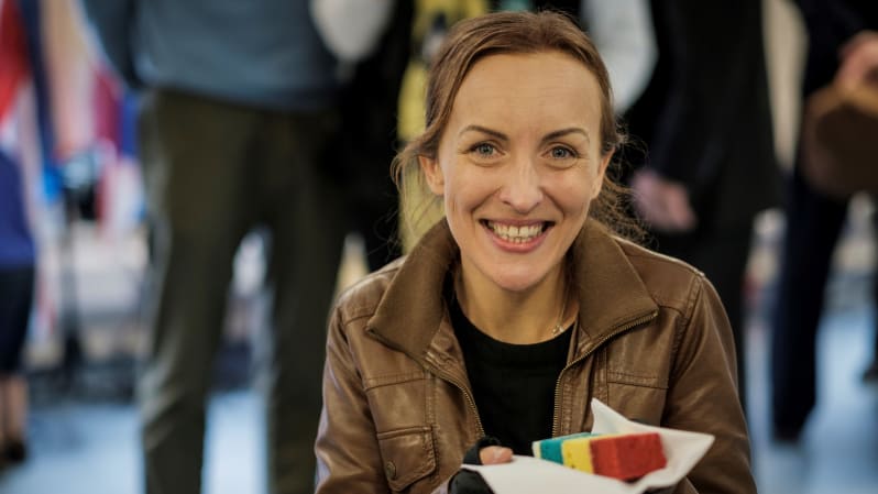Smiling woman in brown leather jacket holds a piece of multicoloured cake in a white napkin. More people blurred in background. 