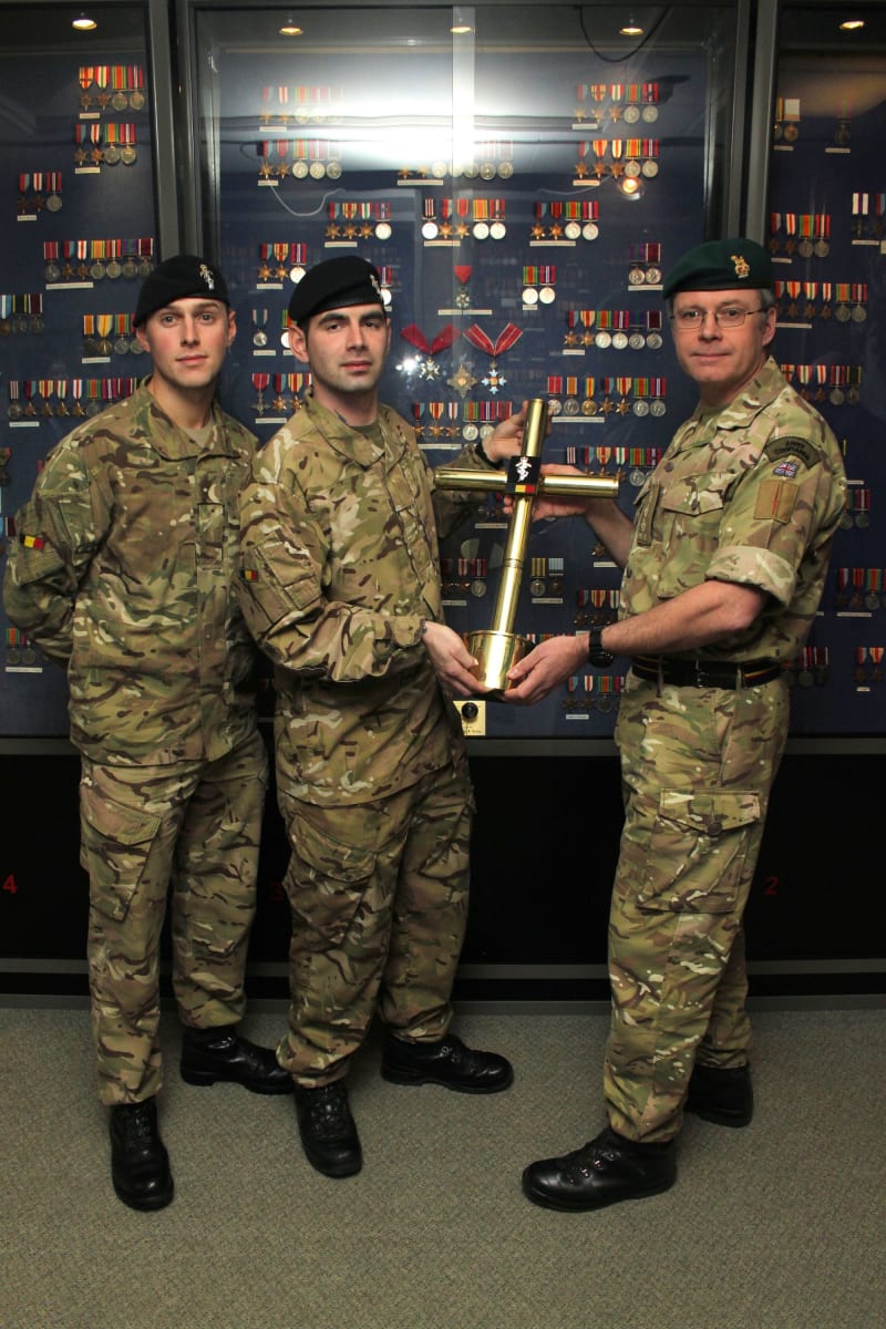 Three soldiers in camouflage uniform. Two on the right hold between them a large gold cross. All look at camera. In the background is a wall of military medals