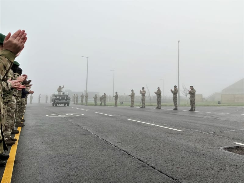 Front-on view of the Land Rover driving in the distance with Col Wigmore stood waving in the back. Soldiers lining the street on either side and clapping. It is a foggy day.