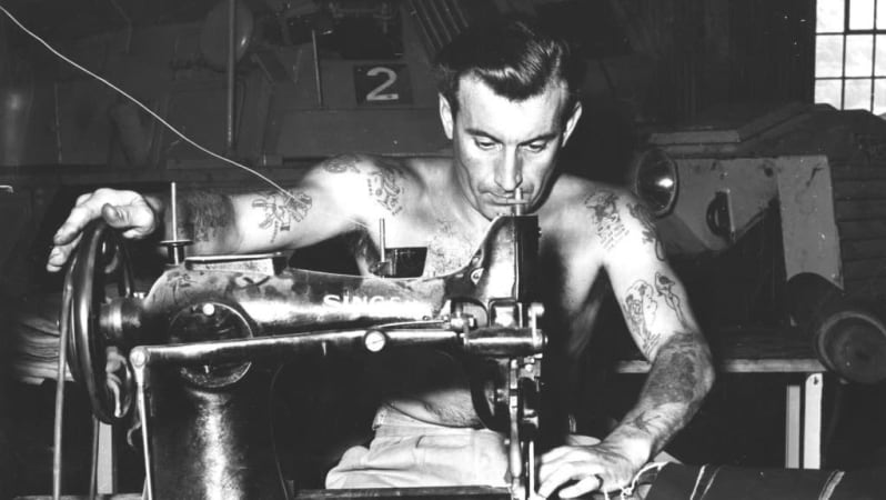 Black and white photo of topless tattooed soldier sat at desk with sewing machine on top. He looks at piece of material under needle. His right hand is on the wheel of the machine.  