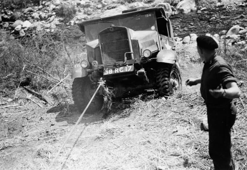 Black and white photograph of a vehicle being pulled out of a river bed. A soldier stands to the side directing.