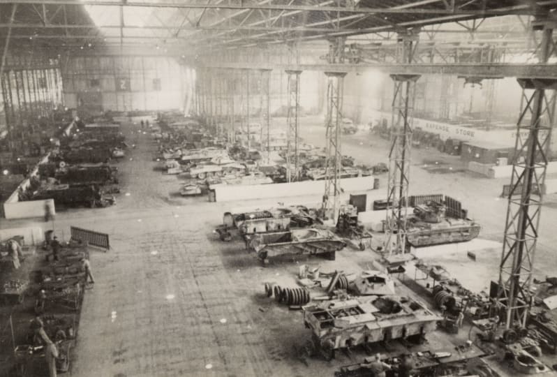 High view of a workshop in a large hangar with several vehicles and tanks lined up inside, black and white.