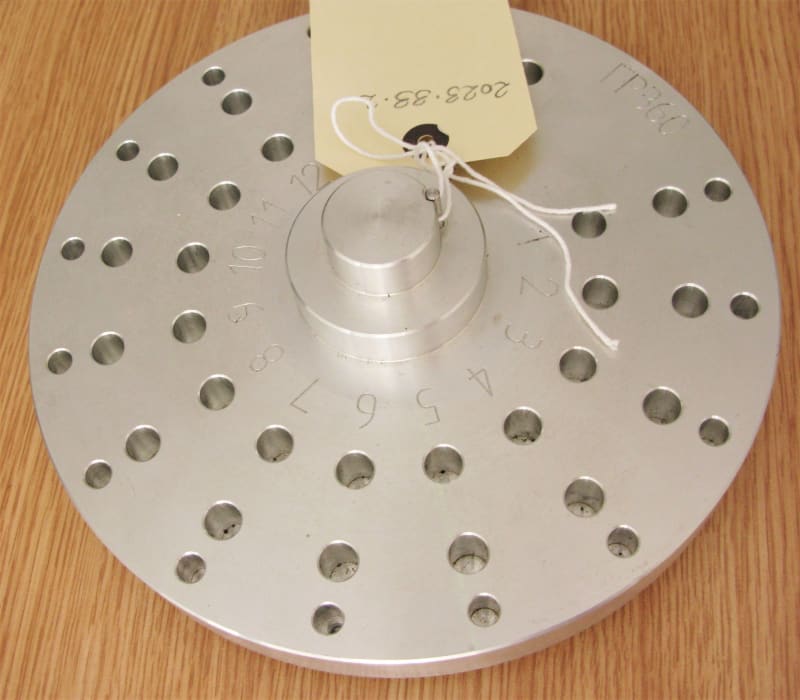 A silver circular piece of metal with holes drilled in circles about the centre.