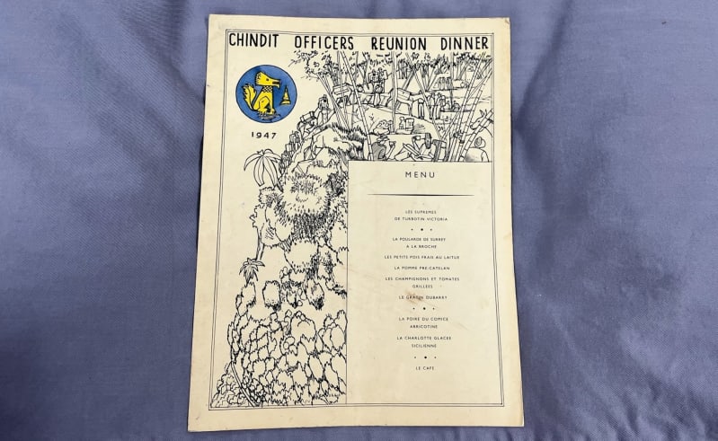A4 piece of card with typed menu, line drawing illustration of troops in the jungle and a stylised lion in yellow, inside a blue circle in the upper-left hand corner. All on a grey blue cushion background. 