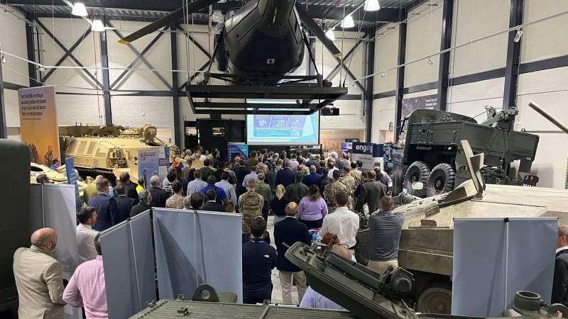 A large hangar gallery with vehicles around the edge and a helicopter hanging from the ceiling. A crowd gathered in the centre of the room, projector screen and pop up banners.