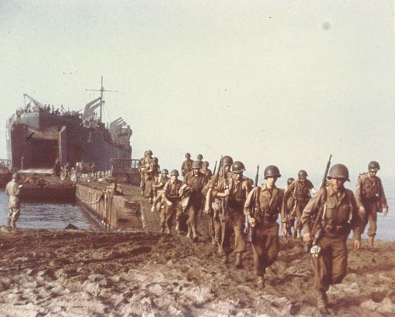 Soldiers disembarking a landing craft at sea onto a beach. 