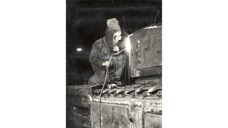 Black and white photograph showing a metalsmith in overalls and welding mask kneeling on an armoured vehicle welding. Sparks are flying.