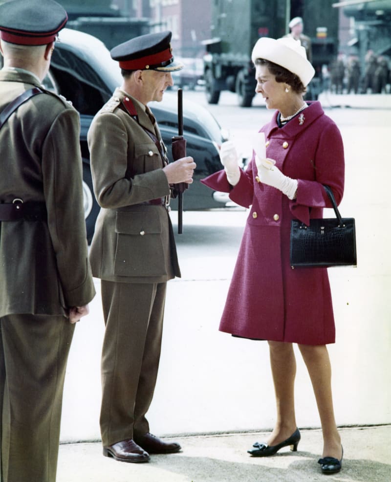 Photograph of Princess Marina wearing a long pink/purple coat with brooch on collar and white hat, stood outside talking to a senior army officer. Another officer looks on with his back to the camera.