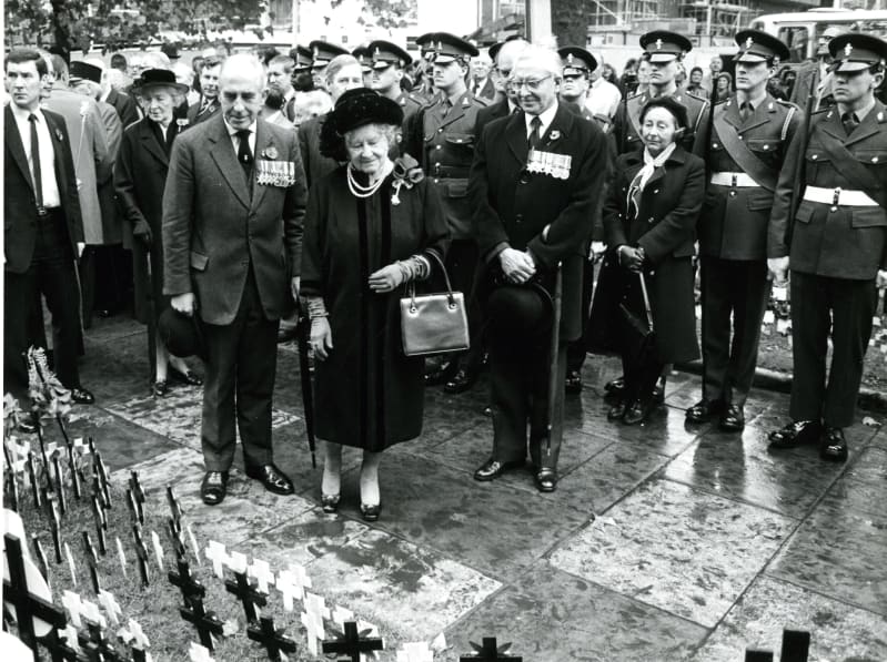 The Queen Mother dressed in black stands looking at Remembrance crosses on the ground. A group of people in military uniform stand behind her.